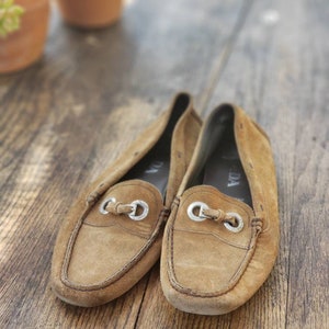 May include: A pair of brown suede loafers with silver buckles. The loafers have a worn look and are sitting on a wooden surface.