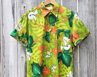 Op de afbeelding: Een groen, kort mouwig shirt met een tropische print met witte, oranje en groene bloemen.