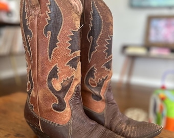 May include: A pair of brown leather cowboy boots with decorative stitching and a pointed toe. The boots show signs of wear, with a distressed appearance and varying shades of brown and tan. They are placed on a wooden surface, suggesting they are vintage.