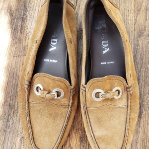 May include: A pair of brown suede loafers with silver metal accents. The loafers have the word "PRADA" printed on the inside of each shoe. The shoes are made in Italy.