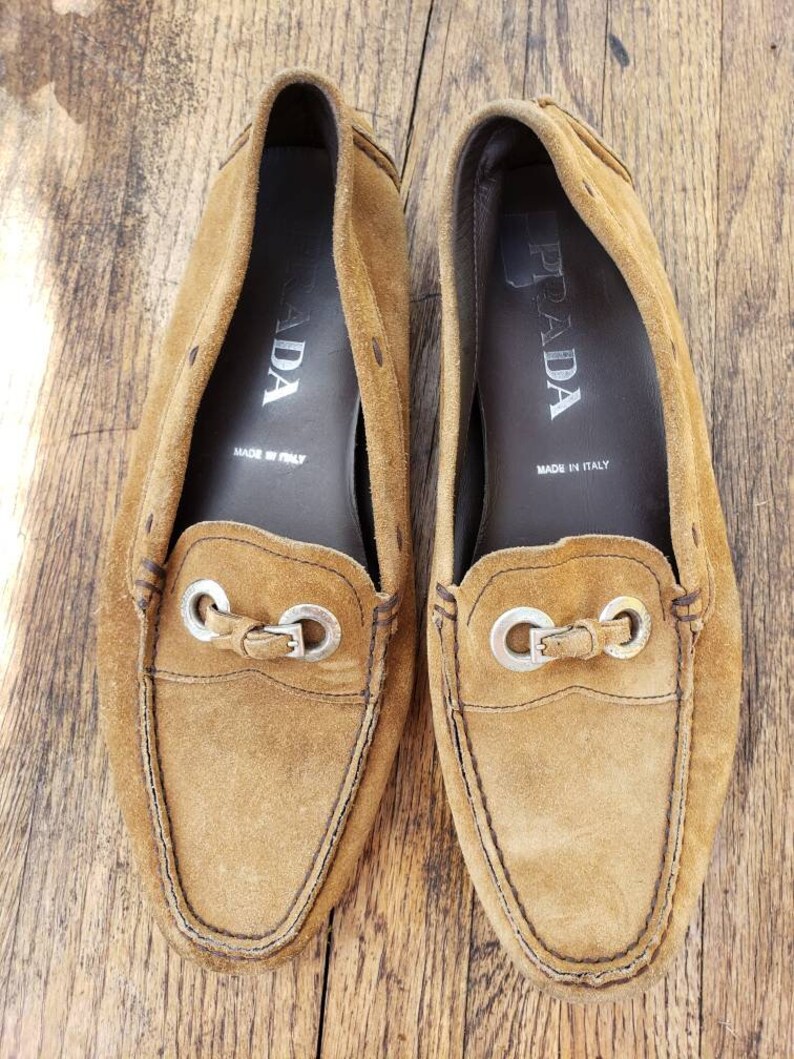 May include: A pair of brown suede loafers with silver metal accents. The loafers have the word "PRADA" printed on the inside of each shoe. The shoes are made in Italy.