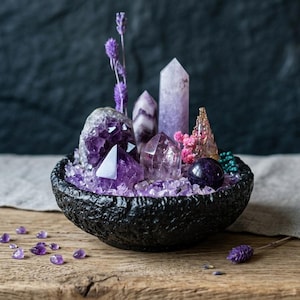 Puede incluir: Un cuenco negro texturizado lleno de cristales de amatista morados, incluyendo varias puntas, un racimo y una esfera lisa. El conjunto está decorado con ramas de lavanda seca y pequeñas flores rosas, con piedras moradas sueltas esparcidas.