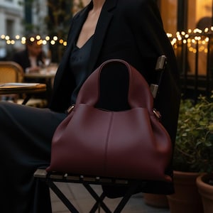 May include: A deep burgundy handbag with a textured surface, featuring a rounded silhouette, a central vertical seam, and integrated top handles. The bag has a structured yet soft form with gold-toned hardware accents at the handle attachments.