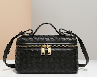 May include: A black rectangular vanity bag featuring a woven leather exterior pattern. The bag includes a top handle, an adjustable shoulder strap, and a gold-toned metal zip closure with two matching pulls centred on the front panel.