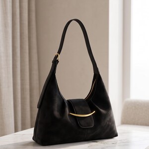 May include: A black shoulder bag with a soft, matte texture, featuring a single adjustable strap with a gold-tone buckle and a central gold-tone curved metal accent on the front flap closure. The bag has a structured yet slouchy silhouette.