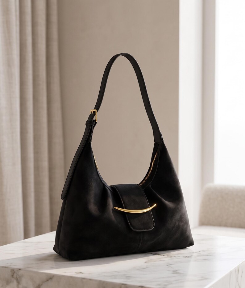 May include: A black shoulder bag with a soft, matte texture, featuring a single adjustable strap with a gold-tone buckle and a central gold-tone curved metal accent on the front flap closure. The bag has a structured yet slouchy silhouette.