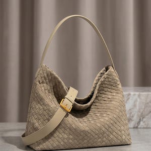 May include: A beige hobo bag featuring an intricate woven leather pattern across the entire exterior. The bag includes a single shoulder strap and a decorative front strap with a gold-toned metal buckle closure, providing a structured yet soft shape.