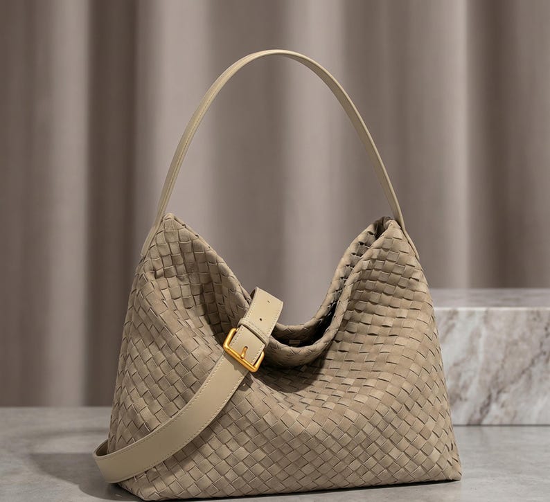 May include: A beige hobo bag featuring an intricate woven leather pattern across the entire exterior. The bag includes a single shoulder strap and a decorative front strap with a gold-toned metal buckle closure, providing a structured yet soft shape.