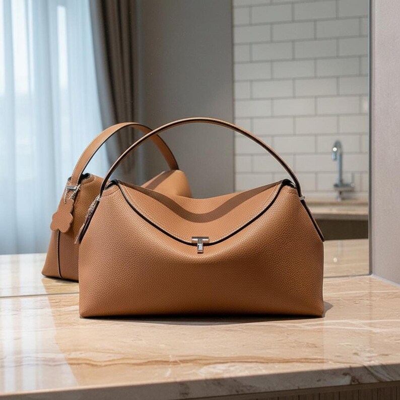 Pair of tan pebbled leather shoulder bags on marble counter, softly structured crescent shape with straps