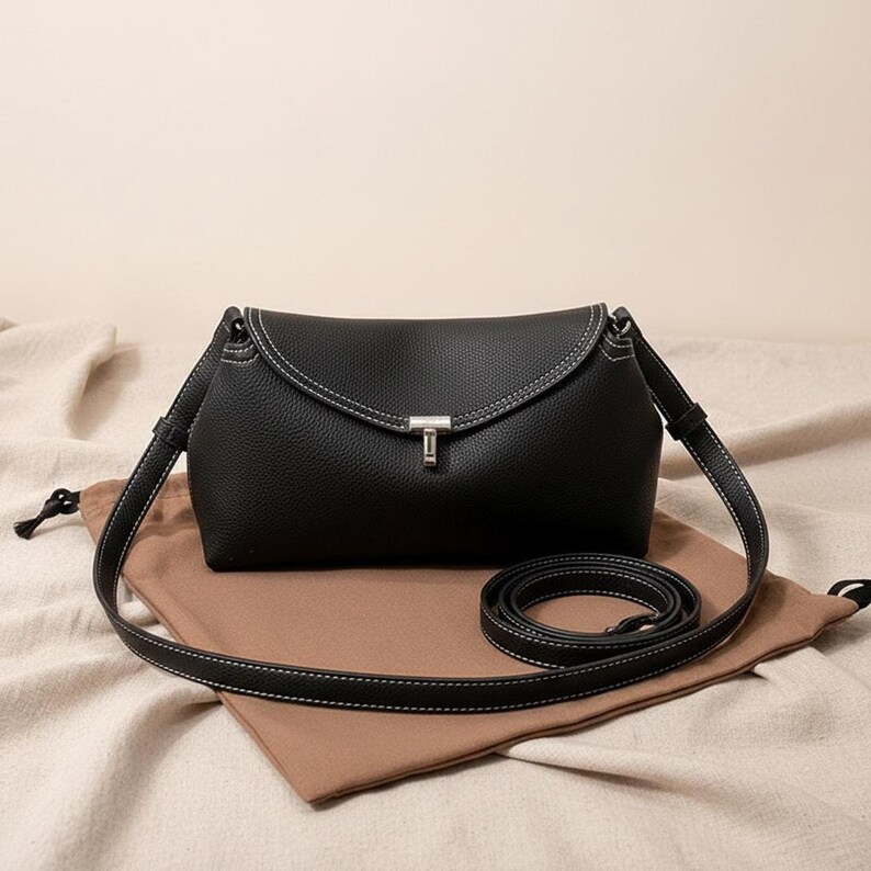 Black pebbled leather shoulder bag on cloth, adjustable strap coiled beside, minimalist crossbody purse