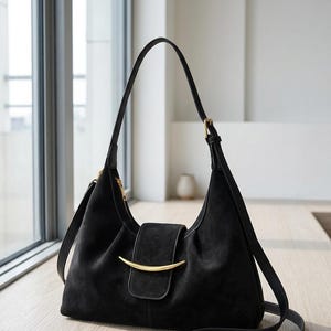 May include: A black shoulder bag with a soft, textured finish, featuring a curved gold-tone metal accent on the front flap closure. The bag has a single adjustable shoulder strap, a top zipper, and a structured, slightly slouchy silhouette.
