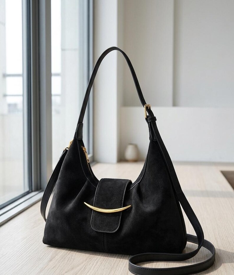 May include: A black shoulder bag with a soft, textured finish, featuring a curved gold-tone metal accent on the front flap closure. The bag has a single adjustable shoulder strap, a top zipper, and a structured, slightly slouchy silhouette.