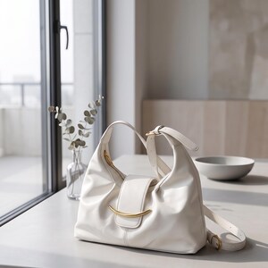May include: A white hobo-style shoulder bag featuring a smooth, soft-touch exterior, a central flap closure secured with a curved gold-tone metal accent, and an adjustable shoulder strap with a matching gold-tone buckle hardware.
