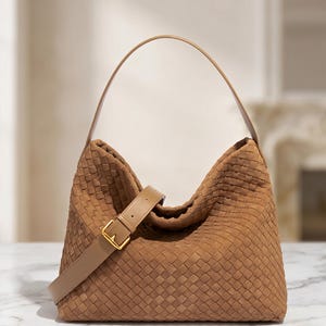 May include: A tan hobo bag featuring an intricate woven leather pattern across the exterior. The bag includes a single arched shoulder strap and an adjustable smooth leather crossbody strap with a gold-toned metal buckle for secure fastening.