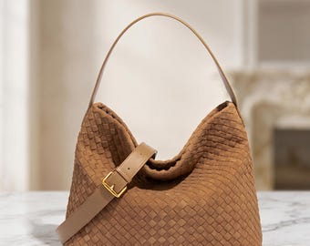 Puede incluir: Un bolso hobo de color tostado con un intrincado patrón de cuero trenzado en el exterior. El bolso incluye una sola correa arqueada para el hombro y una correa cruzada de cuero liso ajustable con una hebilla de metal dorado para el cierre.