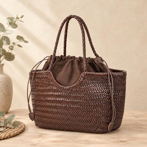 May include: A dark brown woven basket tote bag featuring a herringbone pattern, two braided top handles, and a dark brown fabric drawstring closure insert. The bag has a structured rectangular base and a curved opening at the top.