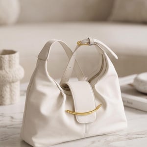May include: A white hobo-style shoulder bag featuring a soft, slouchy silhouette, a central flap closure with a curved gold-tone metal accent, and an adjustable strap with a gold-tone buckle. The bag has a smooth, matte finish.
