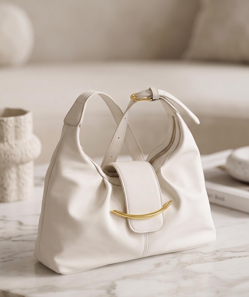 May include: A white hobo-style shoulder bag featuring a soft, slouchy silhouette, a central flap closure with a curved gold-tone metal accent, and an adjustable strap with a gold-tone buckle. The bag has a smooth, matte finish.