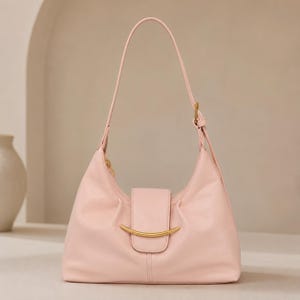 May include: A light pink shoulder bag featuring a smooth, matte finish, a single adjustable strap, and a central flap closure adorned with a curved, gold-toned metal accent piece. The bag has a structured, crescent-shaped silhouette.