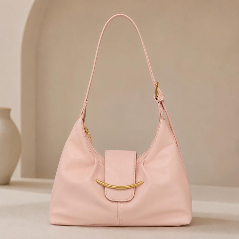 May include: A light pink shoulder bag featuring a smooth, matte finish, a single adjustable strap, and a central flap closure adorned with a curved, gold-toned metal accent piece. The bag has a structured, crescent-shaped silhouette.