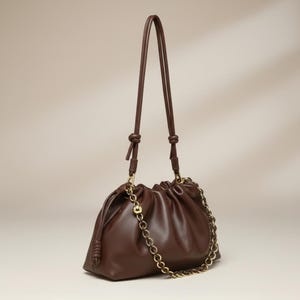 Può includere: Una borsa a tracolla marrone scuro con apertura superiore arricciata. La borsa presenta una tracolla lunga e sottile marrone con dettagli a nodo e una catena decorativa in metallo dorato drappeggiata sul davanti, fissata con elementi dorati.
