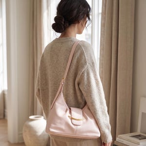May include: A light pink shoulder bag with a smooth finish, featuring a curved gold-tone metal accent on the front flap closure and an adjustable strap. The bag has a structured base and a soft, rounded silhouette, worn over a beige knit sweater.