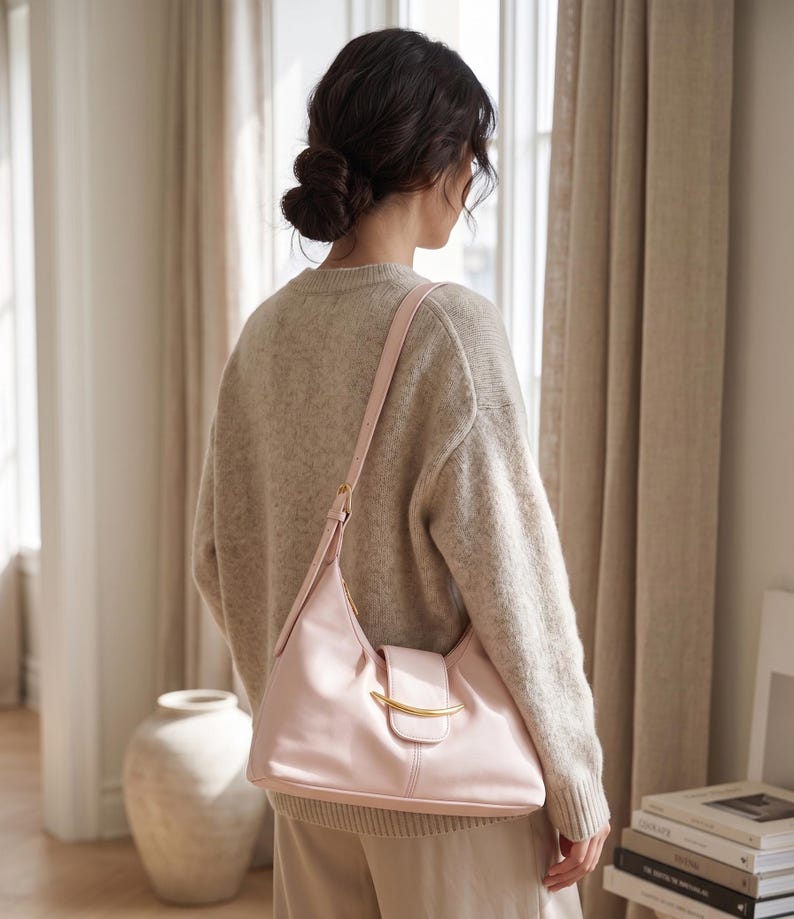 May include: A light pink shoulder bag with a smooth finish, featuring a curved gold-tone metal accent on the front flap closure and an adjustable strap. The bag has a structured base and a soft, rounded silhouette, worn over a beige knit sweater.