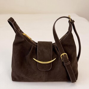 May include: A dark brown suede shoulder bag featuring a gold-toned crescent-shaped metal accent on the front flap. The bag includes a matching adjustable shoulder strap with a gold-toned buckle and a top zipper closure with a gold-toned pull.