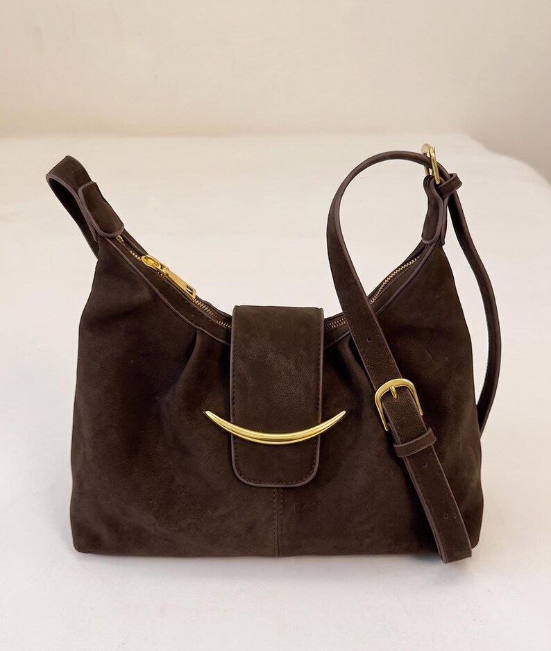 May include: A dark brown suede shoulder bag featuring a gold-toned crescent-shaped metal accent on the front flap. The bag includes a matching adjustable shoulder strap with a gold-toned buckle and a top zipper closure with a gold-toned pull.