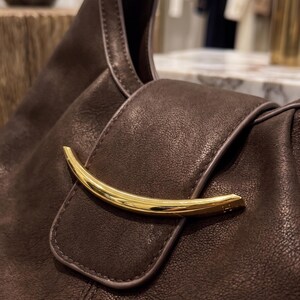 May include: A dark brown leather handbag featuring a textured surface and a prominent, curved, polished gold-tone metal accent piece attached to the front flap closure. The bag has smooth, matching brown piping along the edges of the flap.