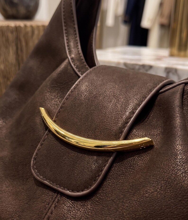 May include: A dark brown leather handbag featuring a textured surface and a prominent, curved, polished gold-tone metal accent piece attached to the front flap closure. The bag has smooth, matching brown piping along the edges of the flap.