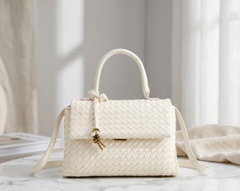 May include: A cream-coloured handbag featuring a woven texture across the exterior. The bag includes a single top handle, a fold-over flap with a knotted cord detail, and a detachable shoulder strap. The design is finished with a small metallic clasp.