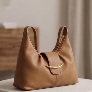 May include: A tan shoulder bag with a smooth texture, featuring a curved gold-tone metal accent on the front flap closure. The bag has two shoulder straps and a gold-tone zipper along the top opening, showcasing a structured silhouette.