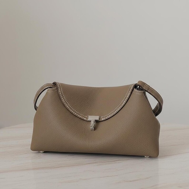 Taupe pebbled leather shoulder bag against gray wall, minimalist crescent silhouette with small metal closure