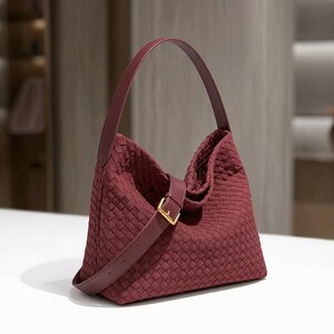 May include: A deep burgundy hobo bag featuring an intricate woven texture across the entire exterior. The bag includes a smooth, solid-coloured shoulder strap and a matching adjustable side strap secured with a polished gold-tone metal buckle.