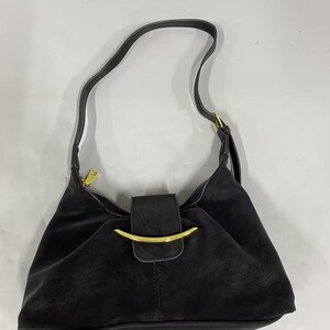 May include: A black shoulder bag featuring a curved gold-tone metal accent on the front flap closure. The bag has a single shoulder strap, a top zipper closure with a gold-tone pull, and a smooth, matte finish throughout the exterior surface.