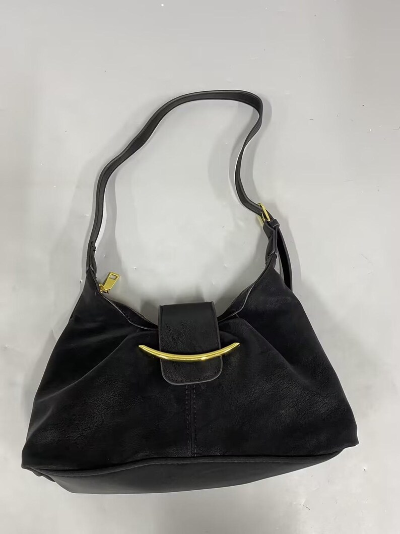 May include: A black shoulder bag featuring a curved gold-tone metal accent on the front flap closure. The bag has a single shoulder strap, a top zipper closure with a gold-tone pull, and a smooth, matte finish throughout the exterior surface.
