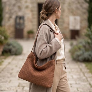 May include: A brown hobo-style shoulder bag featuring a woven leather texture. The bag has a wide, integrated shoulder strap and a slouchy, unstructured silhouette. The exterior surface displays a consistent, intricate basket-weave pattern.