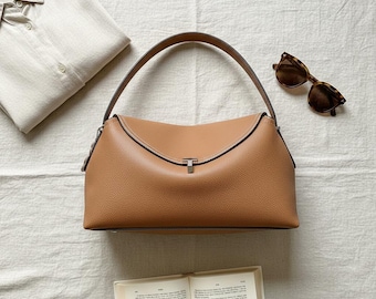 May include: A tan leather handbag with a single arched handle and a central metal clasp closure. The bag features a textured surface and is positioned alongside a light-coloured button-down shirt, a pair of brown tortoiseshell sunglasses, and an open book.