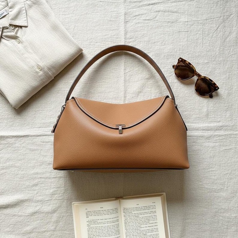 Warm tan pebbled leather shoulder bag on cream linen, curved top handle, metal clasp, sunglasses nearby