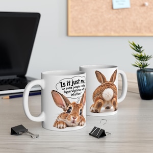 May include: Two white ceramic mugs featuring a brown rabbit. One mug shows the rabbit's face with a thought bubble containing the text: Is it just me... or some people confuse hypervigilance with intuition? The other mug shows the rabbit's rear.