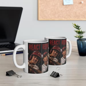 May include: A white ceramic mug featuring a graphic print of a baseball player in a red helmet swinging a bat. The design includes the word REACT in bold red letters above the player. The mug has a handle and a smooth, cylindrical body.