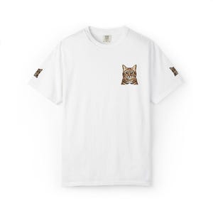 Può includere: Una t-shirt bianca a maniche corte con una piccola illustrazione di un gatto soriano marrone sul lato sinistro del petto. Lo stesso piccolo grafico del gatto soriano marrone è stampato anche sul bordo esterno di entrambe le maniche.