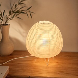 May include: A spherical table lamp featuring a white paper shade with horizontal ribbed detailing. The lamp is supported by a thin, white metal tripod base and emits a warm, soft glow from within, connected by a white power cord.