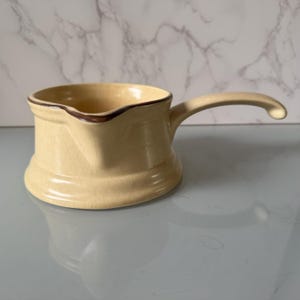 Op de afbeelding: Een crèmekleurige keramische steelpan met één handvat en een schenktuit. De buitenkant is voorzien van een subtiele craquelé glazuurafwerking en een donkerbruin randdetail langs de bovenkant van de pan voor een opvallend contrast.