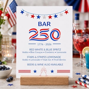 May include: A white rectangular bar menu sign featuring red and blue text and graphics. The sign displays the text BAR 250, 1776-2026, and two cocktail recipes: Red White & Blue Spritz and Stars & Stripes Lemonade, with a wooden base stand.