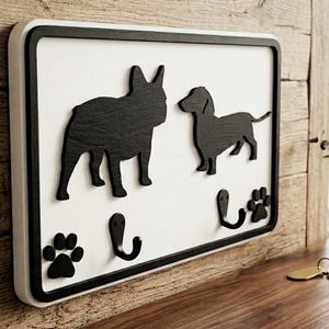 May include: A rectangular wall-mounted key holder featuring a white background with a black border. The design includes two black dog silhouettes, two black metal hooks for hanging items, and two black paw print accents on the white surface.