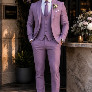 Op de afbeelding: Een driedelig lavendelkleurig pak bestaande uit een getailleerde blazer, een vest met knopen en een bijpassende broek. De outfit bevat een wit overhemd met kraag, een lavendelkleurige stropdas, een wit pochet en donkerbruine leren nette schoenen.