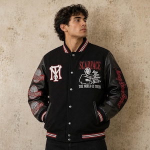 Pode incluir: Uma jaqueta varsity preta com mangas de couro e vários emblemas bordados. A frente exibe o logotipo Scarface, um gráfico de personagem e o texto The World Is Yours. As mangas apresentam ícones de globo e o texto Money Power Respect.