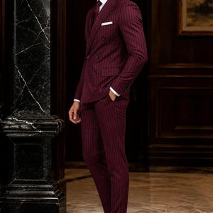 Op de afbeelding: Een double-breasted pak in donkerbordeaux met dunne witte krijtstrepen, inclusief een wit overhemd, een donkerrode das, een wit pochet en donkerbruine leren nette schoenen. Het colbert heeft puntrevers en zwarte knopen.